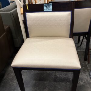 Beige cushioned wooden chair on display in a store, with other chairs and caution tape in the background.
