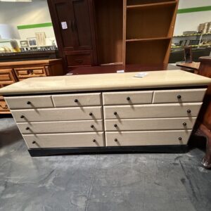 A beige dresser with eight drawers and black knobs is displayed in a furniture store.