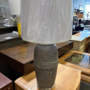 A textured table lamp with a white shade wrapped in plastic sits on a wooden display in a store.