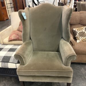 Olive green velvet wingback armchair with dark wood legs in a furniture store setting.