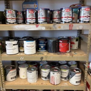 Cans of paint in various colors and brands arranged on metal shelves in a storage area.