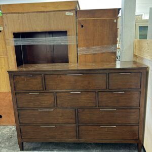 A wooden dresser with ten drawers and metal handles stands in a warehouse next to other furniture.