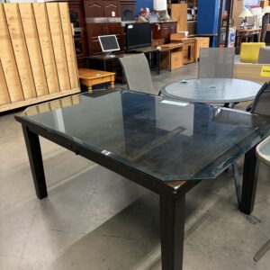 Rectangular glass-top table with black legs in a store, surrounded by other furniture on a concrete floor.