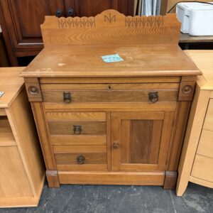 Antique wooden dresser with a carved backboard, three drawers, and a single cabinet door on a concrete floor.