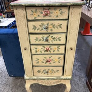 Cream-colored vintage cabinet with floral designs and five drawers, standing on curved legs in a store.