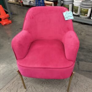 Bright pink armchair with gold legs, sitting on a rug in a warehouse with paint cans and shelves in the background.