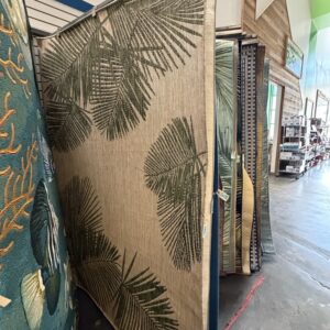 Large beige rug with green palm leaf pattern displayed in a store aisle among other rugs.