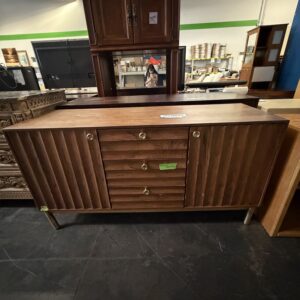 Mid-century modern wooden credenza with vertical grooves and brass handles in a thrift store setting.