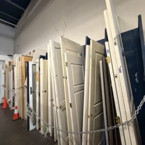 A row of used doors leaning against a wall, with two orange traffic cones and a chain blocking entry.