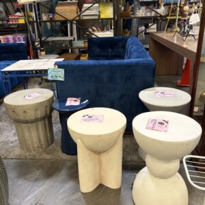Four modern stools with price tags sit in front of a blue couch in a thrift store.