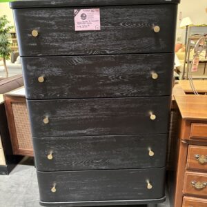 Tall black wooden dresser with five drawers, gold knobs, and tapered legs, displayed in a furniture store.