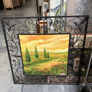 Ornate metal frame with a painting of trees, fields, and flowers, displayed in a warehouse or thrift store.