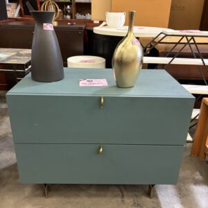 A teal two-drawer chest with gold handles, topped with a black vase and a gold vase, in a store.