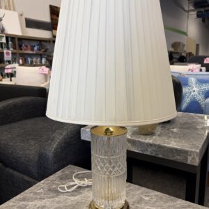 A glass and brass table lamp with a white shade sits on a gray marble table in a furniture store.