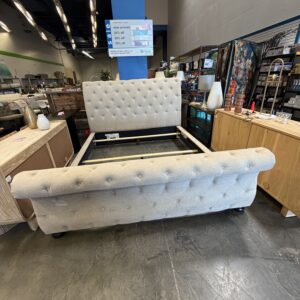 Gray upholstered bed frame on display in a store, surrounded by wooden furniture and home decor items.