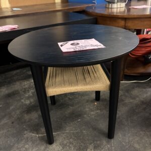 Round black side table with a woven shelf underneath, displayed in a store with other furniture.
