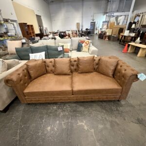 Brown tufted sofa with three cushions displayed in a warehouse with other furniture items.