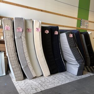 Nine mattresses standing upright in a row on a rug inside a warehouse-like store, each with a pink price tag.