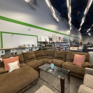 Brown sectional sofa with pillows, glass coffee table, and shelves in a furniture store with bright lighting.