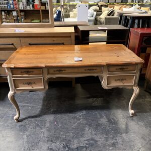 Vintage wooden desk with curved legs and five drawers, displayed in a thrift store with other furniture.