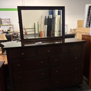 Dark wood dresser with attached mirror, featuring nine drawers, in a warehouse or store setting.