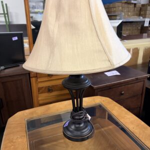 A beige lampshade on a dark metal lamp sits on a glass-topped wooden table in a thrift store.