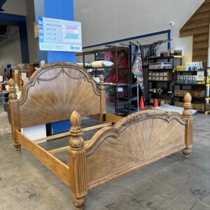 Ornate wooden bed frame with carved posts displayed in a warehouse store with shelves and signage.