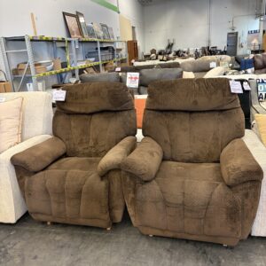 Two brown recliner chairs side by side in a thrift store with other furniture in the background.