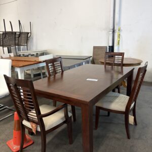 Four wooden chairs around a rectangular wooden table in a warehouse, with stacked chairs and an orange cone nearby.