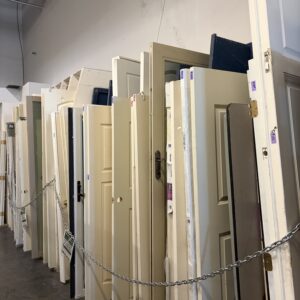 A row of used doors in various colors and styles standing upright in a warehouse.