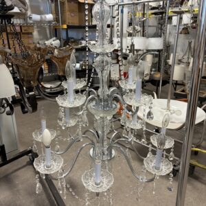 A glass chandelier with multiple arms and bulbs hangs in a warehouse filled with other lighting fixtures.