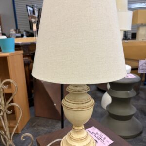 Table lamp with a cream-colored tapered shade and a distressed, pedestal-style base.
