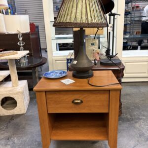 A brown wooden nightstand topped with a table lamp featuring a green and brown wicker-style shade.